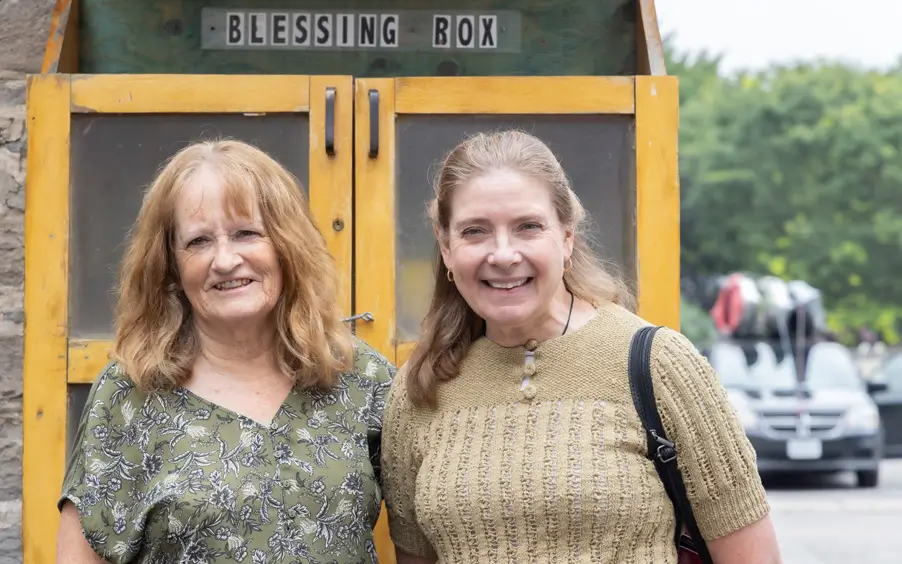 Two women looking at the camera posing on front of a wooden cabinet outside of the Anglican Church in Stirling, ON